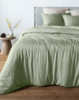 A bed dressed in a sage green bambaw cozy bamboo duvet cover and matching pillowcases, styled in a bedroom with a wooden floor, a light gray headboard, and a small wooden side table with a vase of branches.