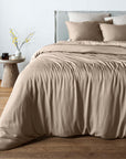 A bed dressed in a bambaw cozy bamboo taupe beige duvet cover and matching pillow shams, styled in a minimalist bedroom with a wooden floor, a light gray headboard, and a small wooden side table with a vase of yellow branches.