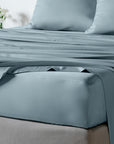 A close-up of bambaw cozy dusty blue fitted bed sheet dressed in a smooth, sheet set with matching pillowcases and a fitted sheet draped over the corner.