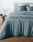 A bed dressed in a dusty blue bamboo duvet cover and matching pillow shams, styled in a bedroom with a wooden floor, a light wood side table, and neutral wall tones.
