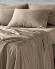 A neatly made bed with taupe-beige bamboo bedding, including a fitted sheet, matching pillowcases, and two large pillows, shown in a softly lit bedroom setting.