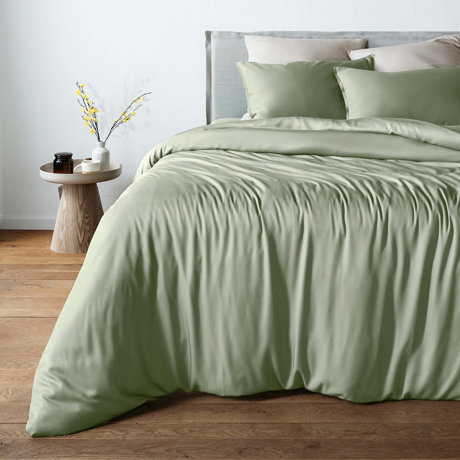 A bed dressed in a sage green bambaw cozy bamboo duvet cover and matching pillowcases, styled in a bedroom with a wooden floor, a light gray headboard, and a small wooden side table with a vase of branches.