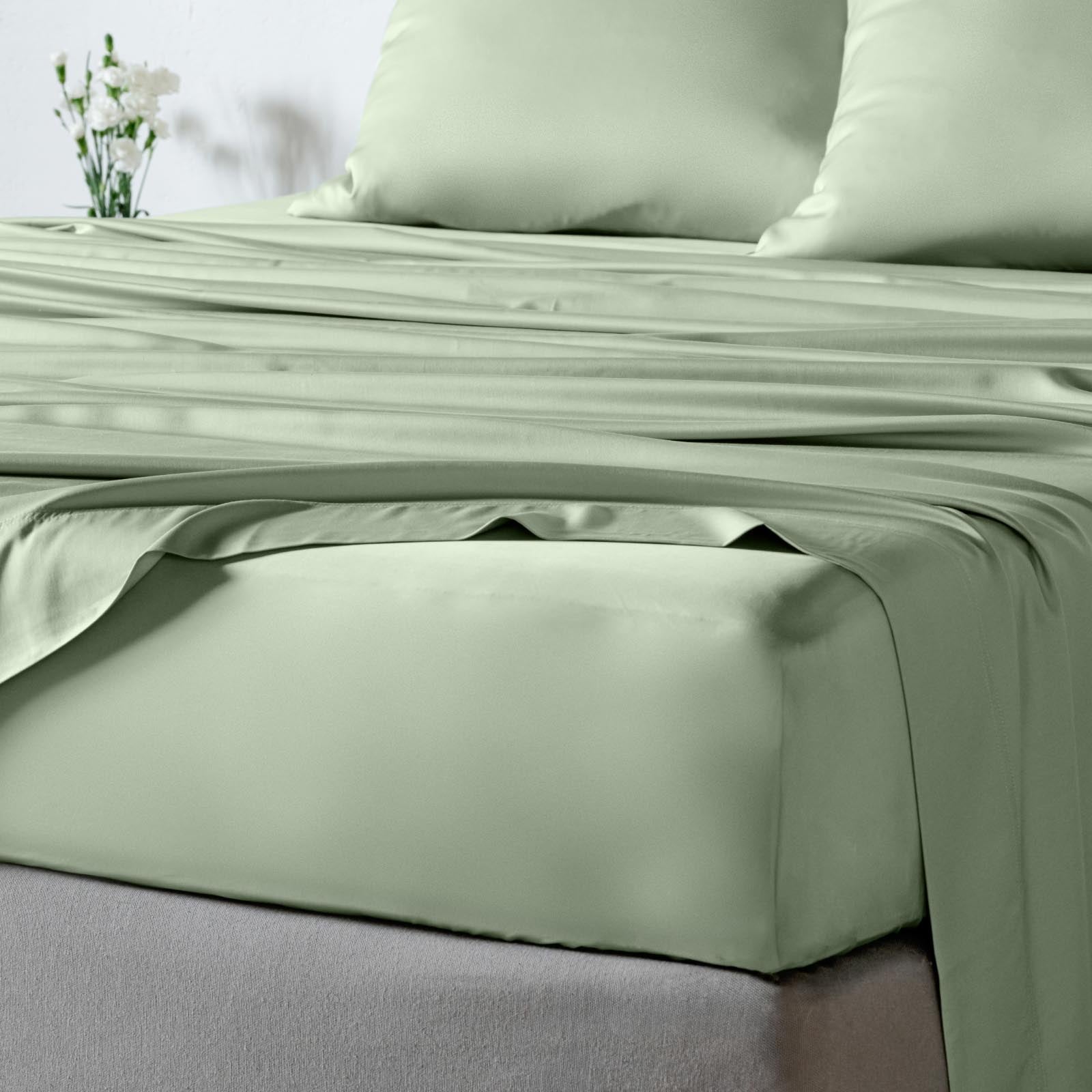 A bed dressed in a smooth, bamboo sage green sheet set with a fitted sheet and matching pillowcases, shown in a softly lit bedroom setting.