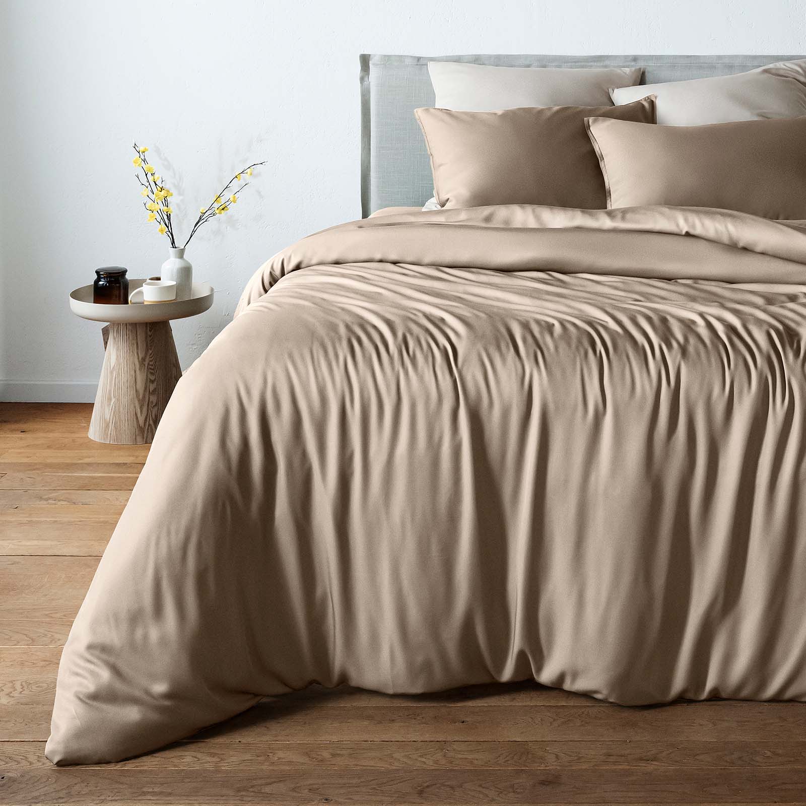 A bed dressed in a bambaw cozy bamboo taupe beige duvet cover and matching pillow shams, styled in a minimalist bedroom with a wooden floor, a light gray headboard, and a small wooden side table with a vase of yellow branches.