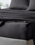 Black bambaw cozy bamboo bed sheet set with a fitted sheet and matching pillowcases on a bed, with a small vase of white flowers in the background.