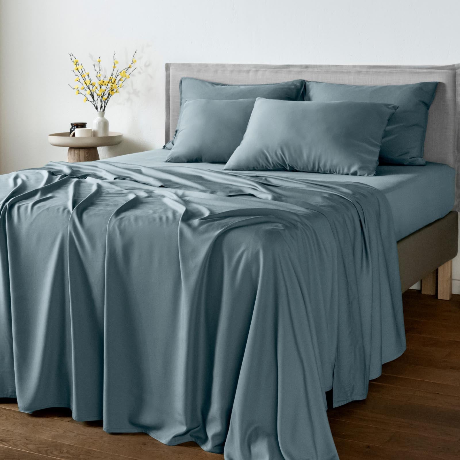 A neatly made bed with bambaw cozy dusty blue bamboo bedding, including a duvet cover and matching pillowcases, styled in a minimalist bedroom setting with a wooden floor and a side table.