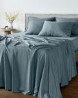 A neatly made bed with bambaw cozy dusty blue bamboo bedding, including a duvet cover and matching pillowcases, styled in a minimalist bedroom setting with a wooden floor and a side table.