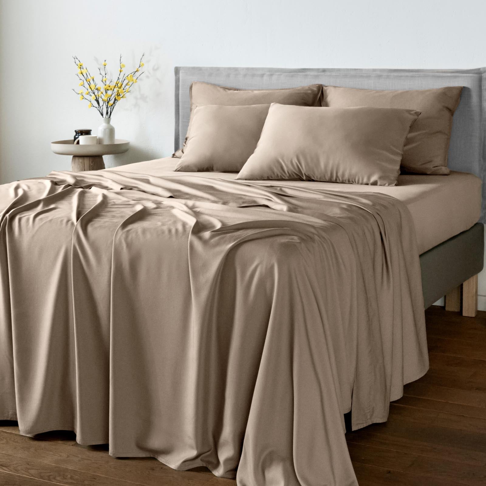 A neatly made bed with a smooth taupe-beige bamboo bedsheet set, including a fitted sheet and matching pillowcases, styled in a modern bedroom setting with a gray upholstered headboard and a small side table with yellow flowers.