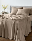 A neatly made bed with a smooth taupe-beige bamboo bedsheet set, including a fitted sheet and matching pillowcases, styled in a modern bedroom setting with a gray upholstered headboard and a small side table with yellow flowers.