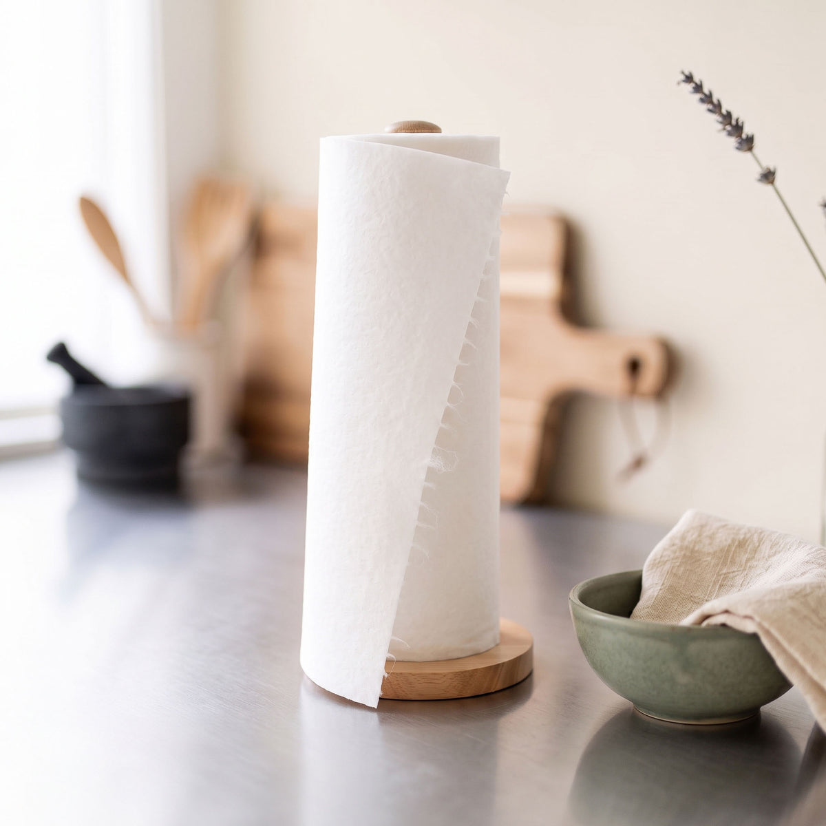 A roll of reusable white paper towels on a wooden stand on a kitchen counter, with a green bowl and wooden cutting boards blurred in the background.