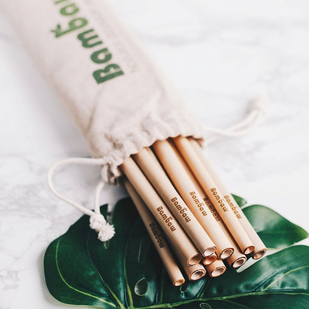 Natural bamboo drinking straws in a drawstring bag placed on a marble surface with green leaves